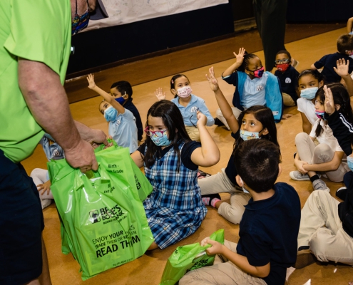 children raise their hands while an adult hands out green Bernie's Book Bank bags