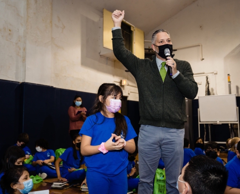 a speaker holds a microphone and raises his hand, next to children wearing surgical masks