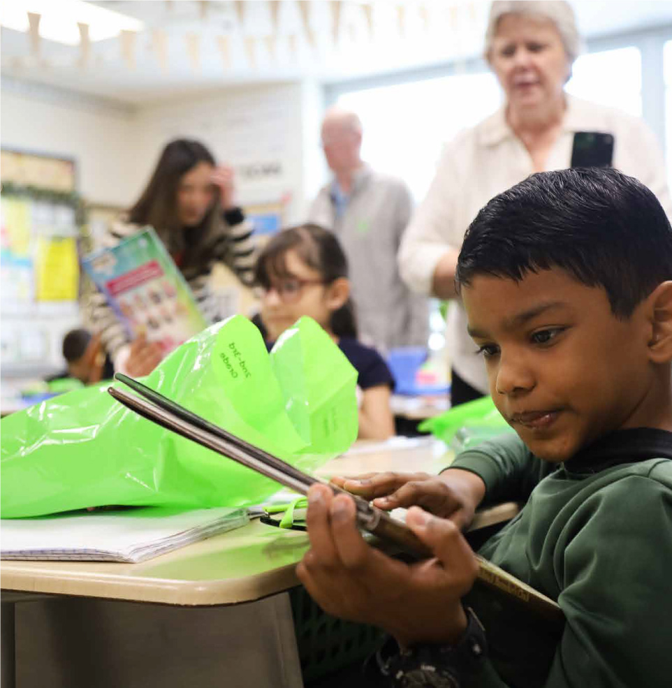 a kid looks at a large picture book with a teacher and classmates visible in the background