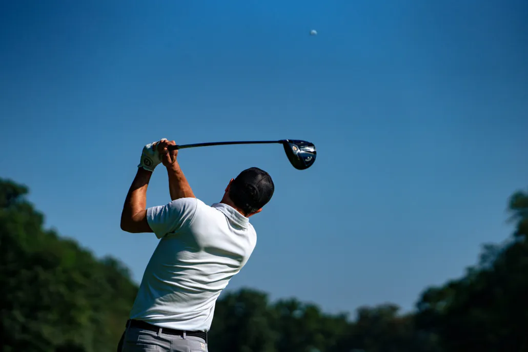 a male golfer takes a massive swing at a golf ball, visible high in the sky