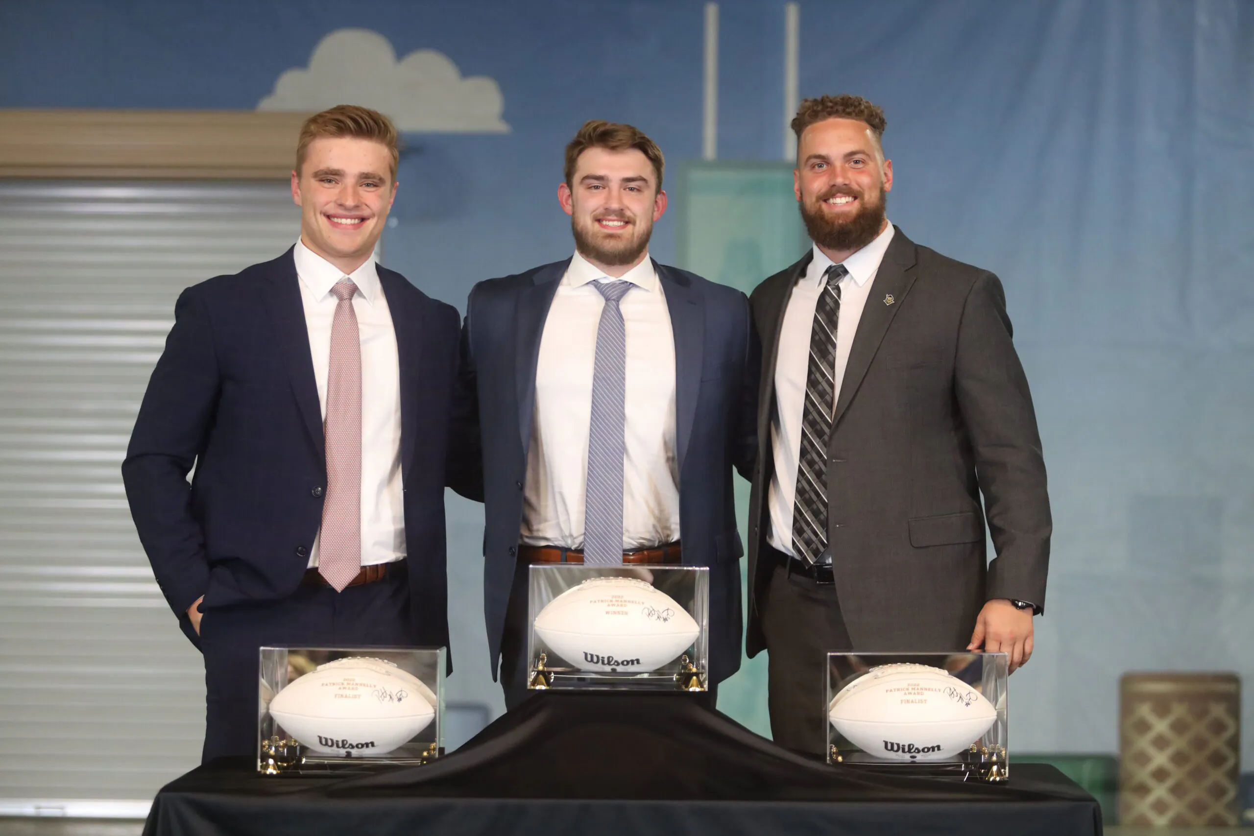 three tall smiling men stand in front of signed footballs
