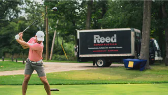 a male golfer prepares a drive with a Reed Construction trailer behind him