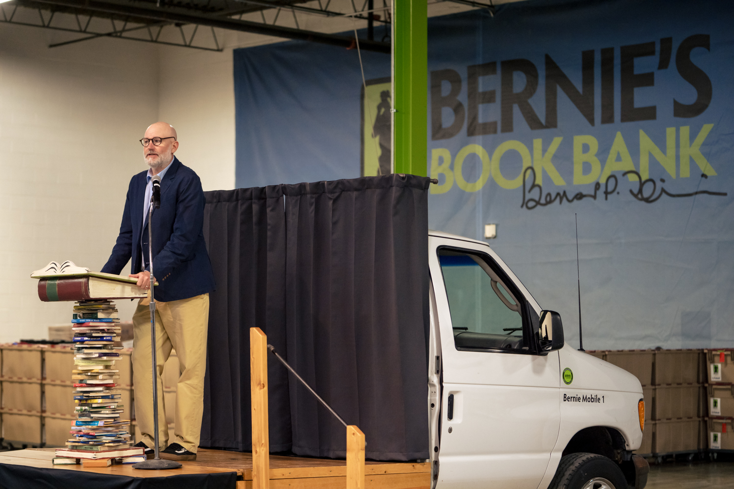 a speaker at a podium in front of a large wall hanging that reads Bernie's Book Bank