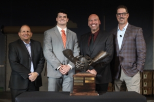 four men wearing suits pose in front of the Patrick Mannelly award, a statue of two bronzed hands that clasp a football