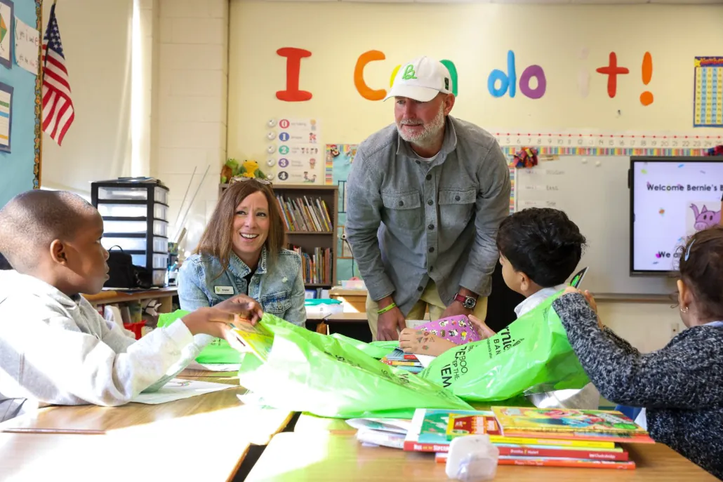two adults in a classroom hand pass out books to a schoolchild