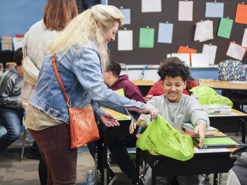 An adult volunteer smiles and hands a middle school-aged kid a green bag of Bernie's books