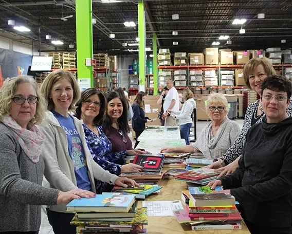 Eight adult women volunteers at Bernie's Book Bank Milwaukee office smile while sorting books