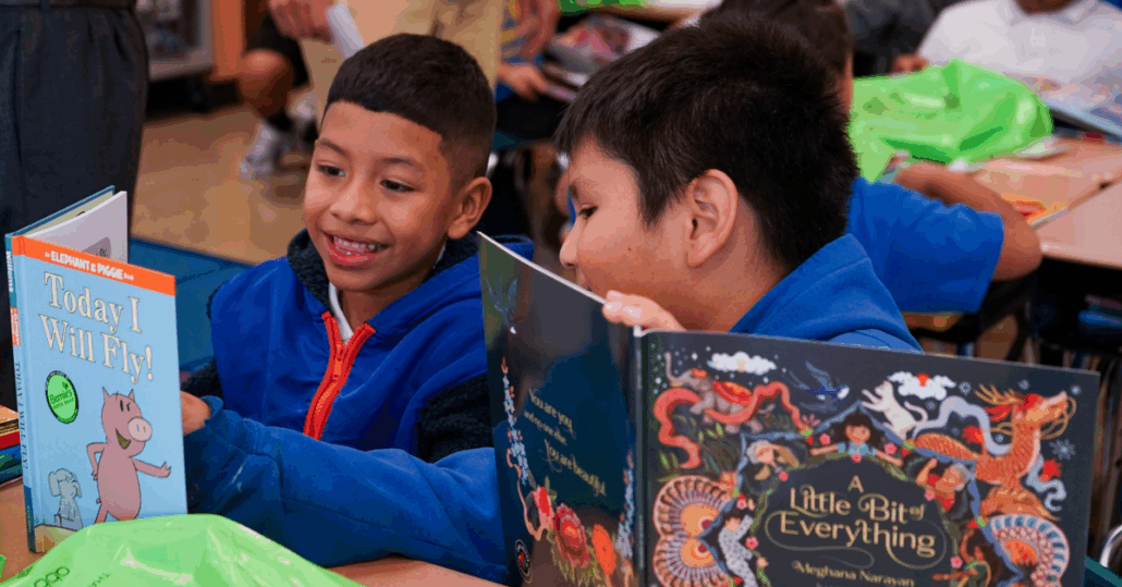 two schoolchildren smile while looking over their books, including Mo Willems's Today I Will Fly with Elephant and Piggie, and A Little Bit of Everything by Meghana Naravan